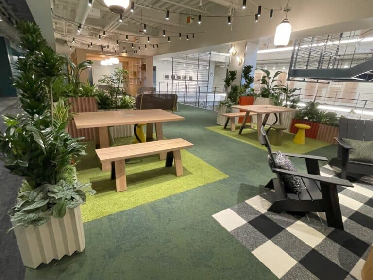 A contemporary office space featuring a large, designer picnic-style table with benches, surrounded by abundant greenery in planter boxes, blending functional workspace with biophilic elements.