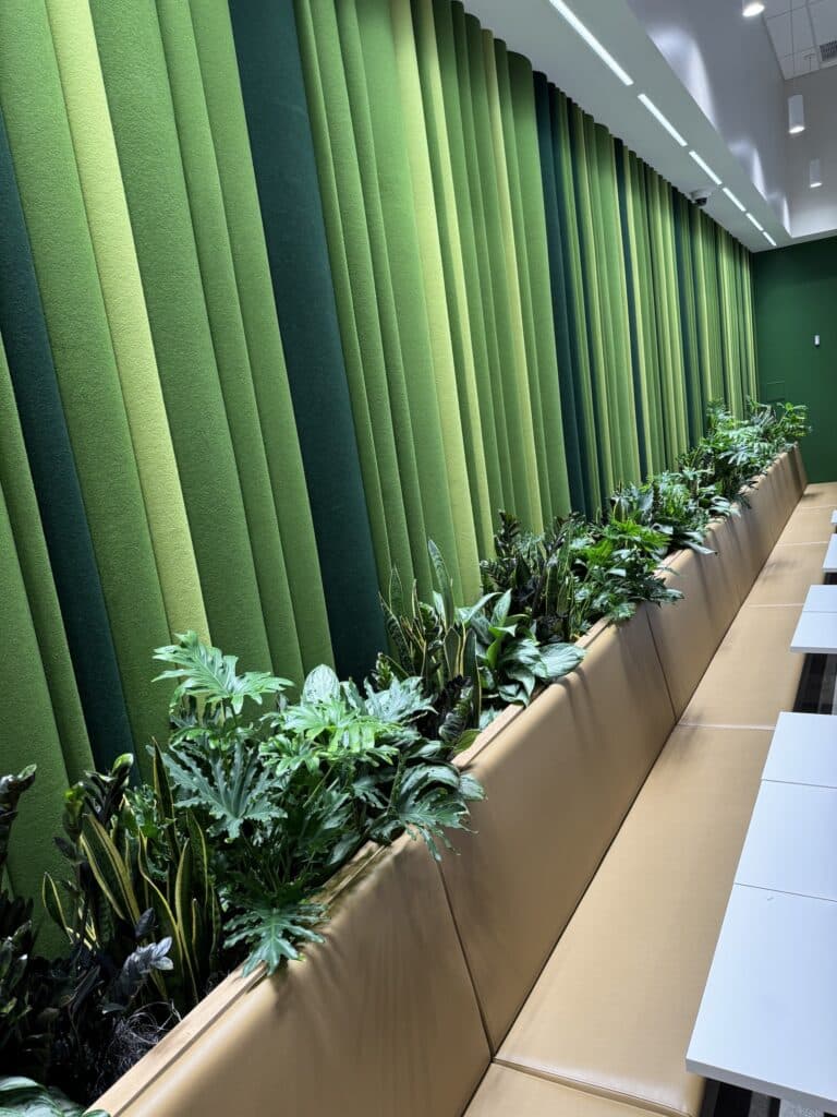Built-in linear planter with dense green foliage along a tan booth, against a fabric-panel green feature wall.